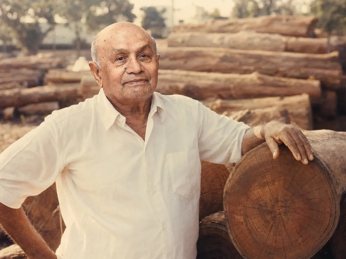 Pachan Dhanji Patel standing beside stacked teak logs at the depot